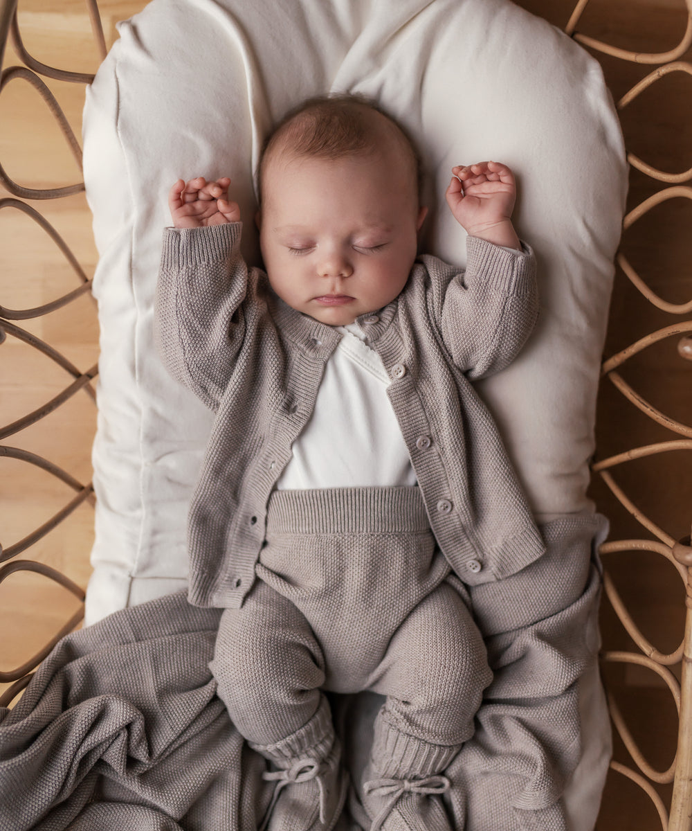 Baby iført beige/grå strikket sæt liggende i flettet kurv med beige tæppe