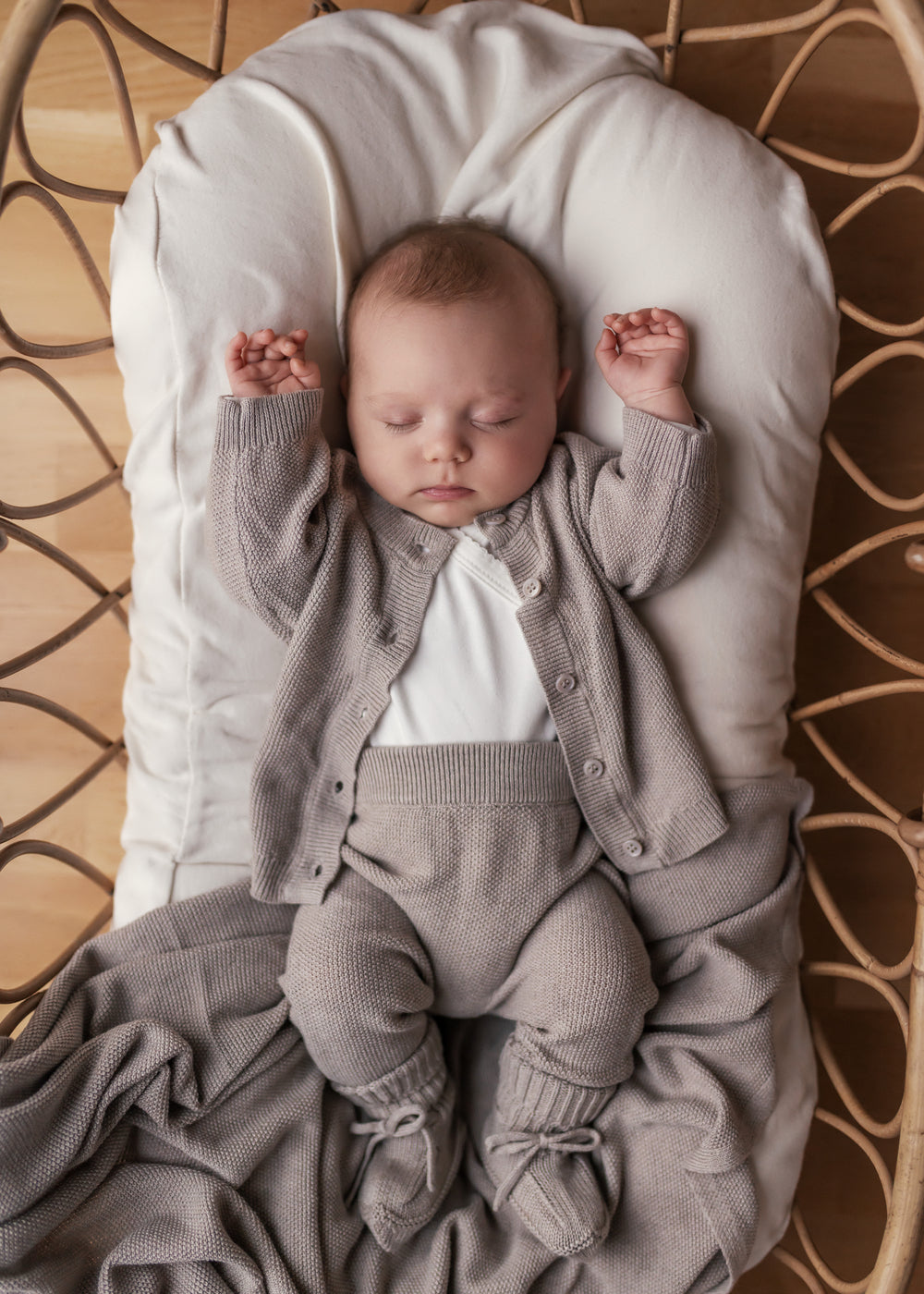 Baby iført beige/grå strikket sæt liggende i flettet kurv med beige tæppe