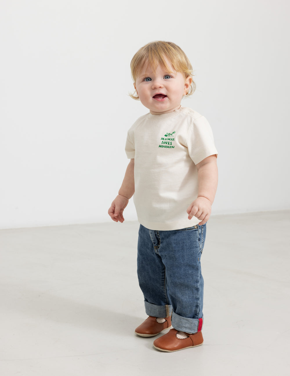 Baby wearing a white T-shirt with a green Moomin print and jeans.