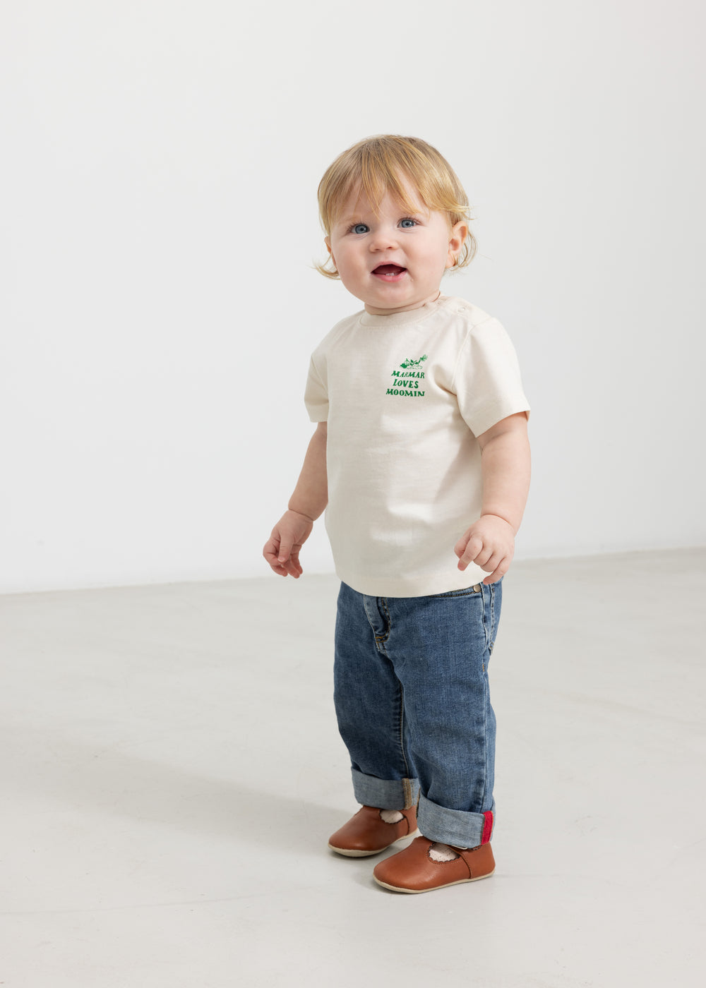 Baby wearing a white T-shirt with a green Moomin print and jeans.