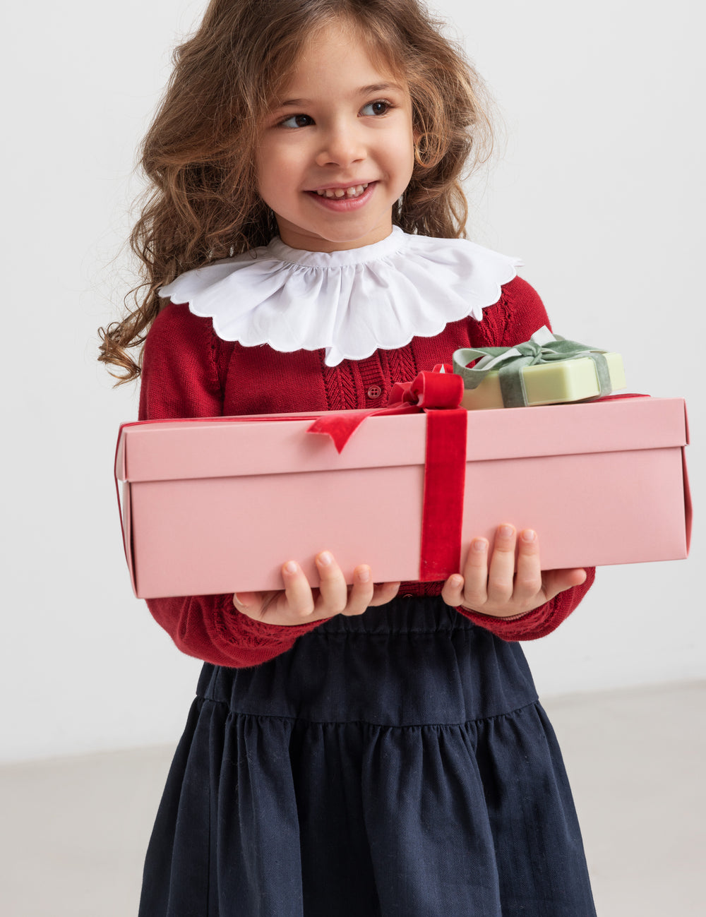 Girl wearing red Christmas sweater with lace pattern and white collar and blue skirt, holding Christmas presents