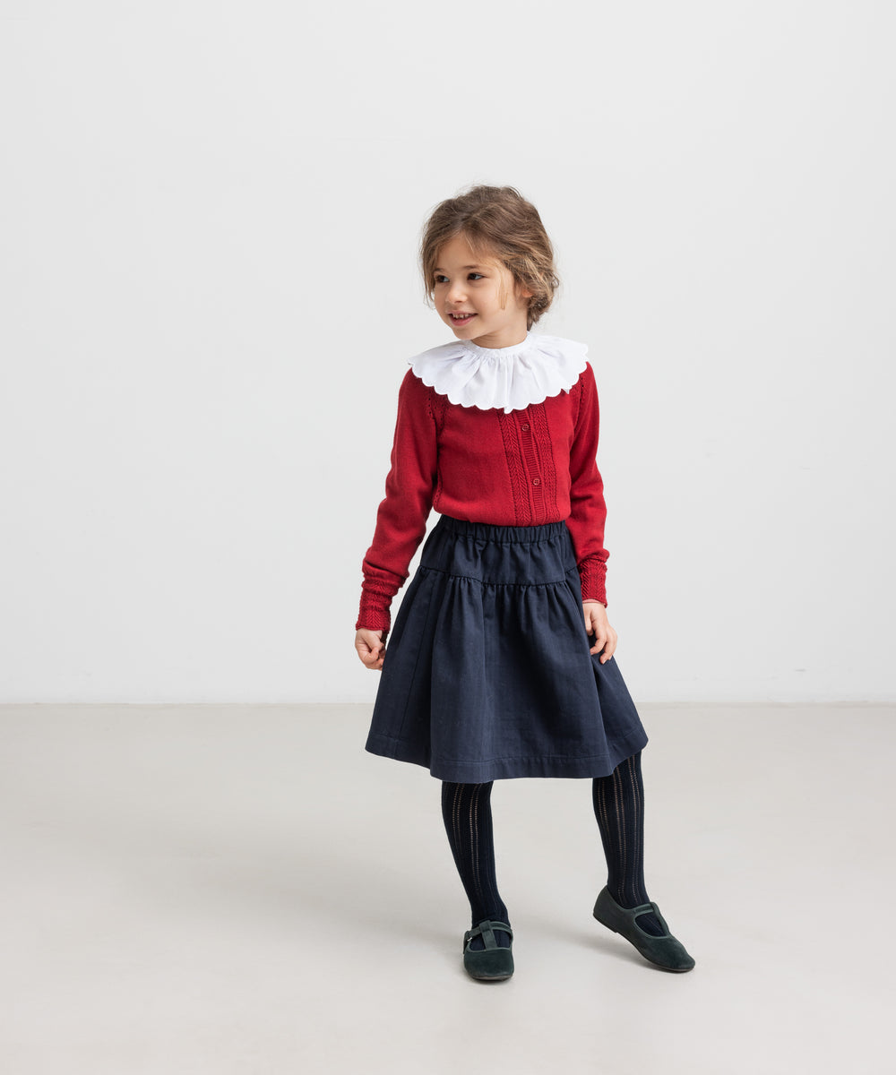 Pige iført rød juletrøje med hulmønster, hvid kravebluse og mørkeblå festnederdel til børn