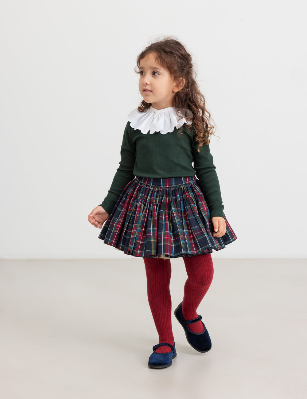 Pige iført juletøj - mørkeblåt og rødt ternet nederdel, mørkegrøn bluse og røde strømpebukser til børn
