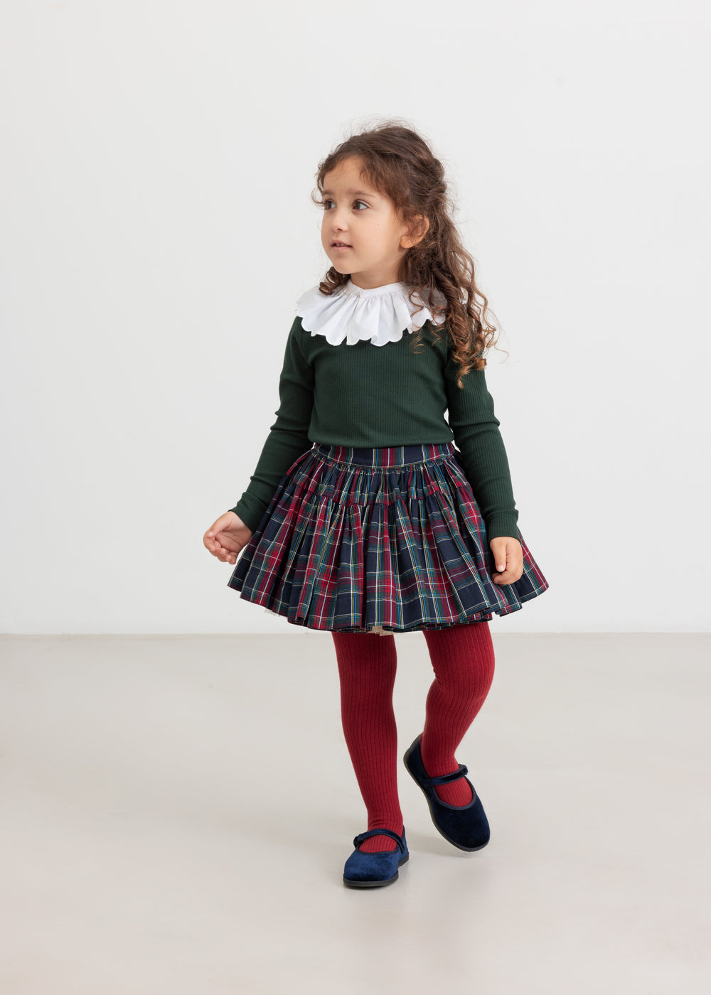 Pige iført mørkegrøn bluse, ternet nederdel og røde strømpebukser – festligt outfit til børn