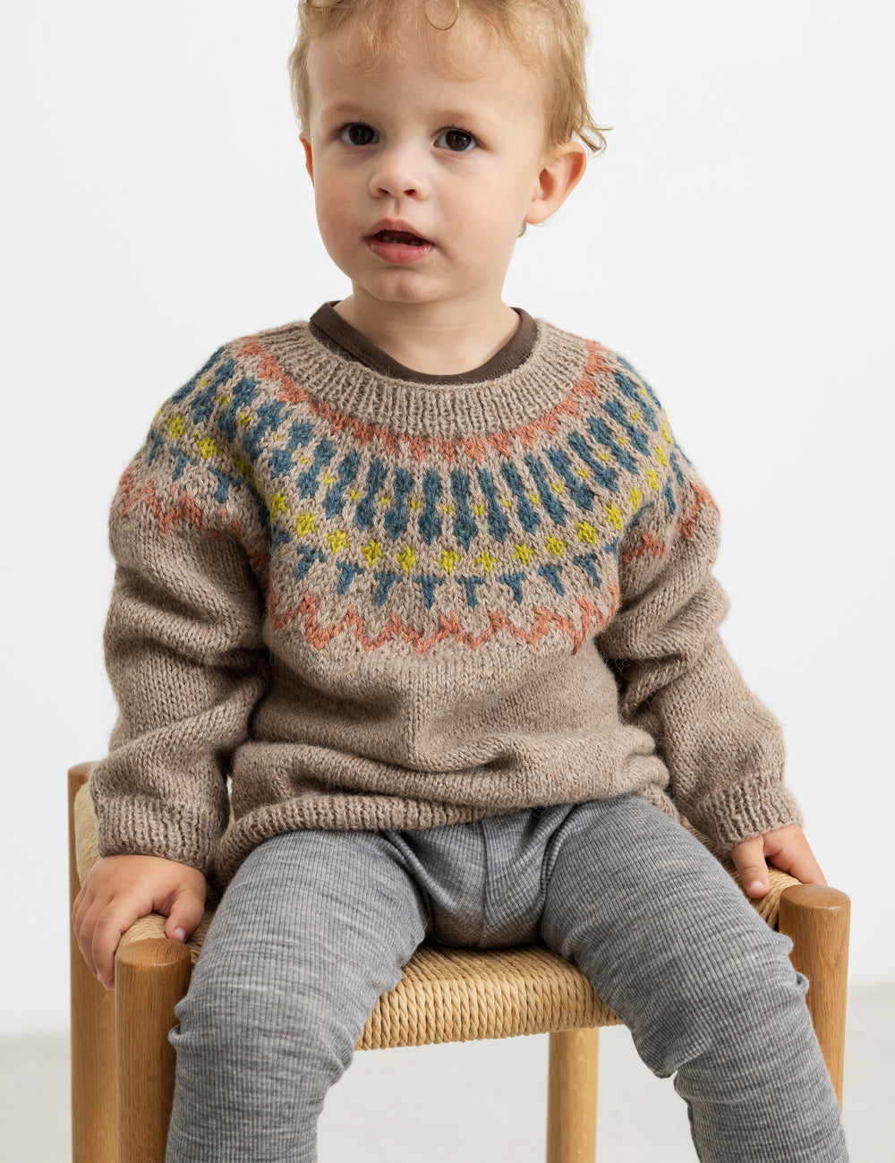 Dreng iført beige uldstrikbluse med mønster sidder på stol.