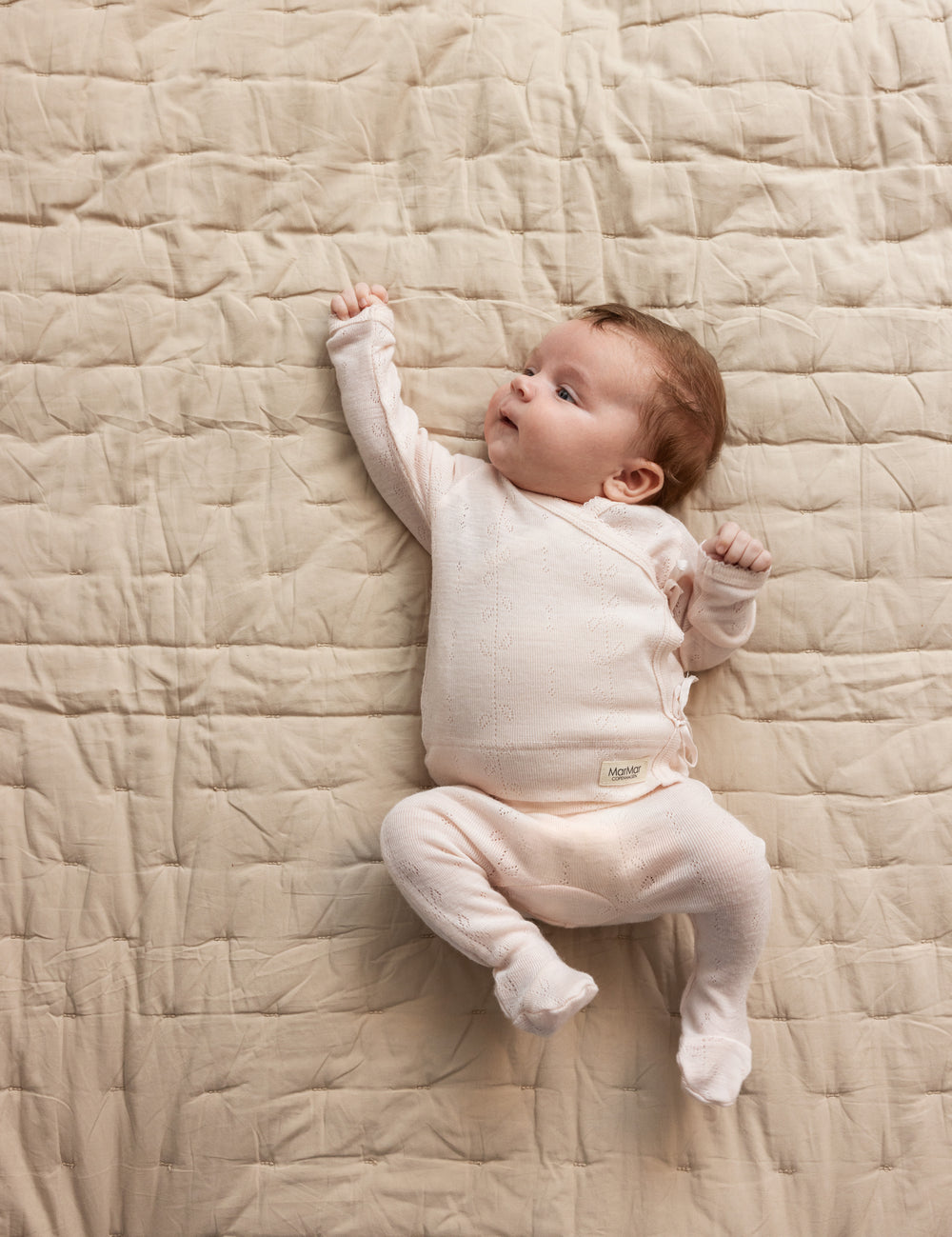 Baby iført lyserød heldragt i merinoud ligger på tæppe.