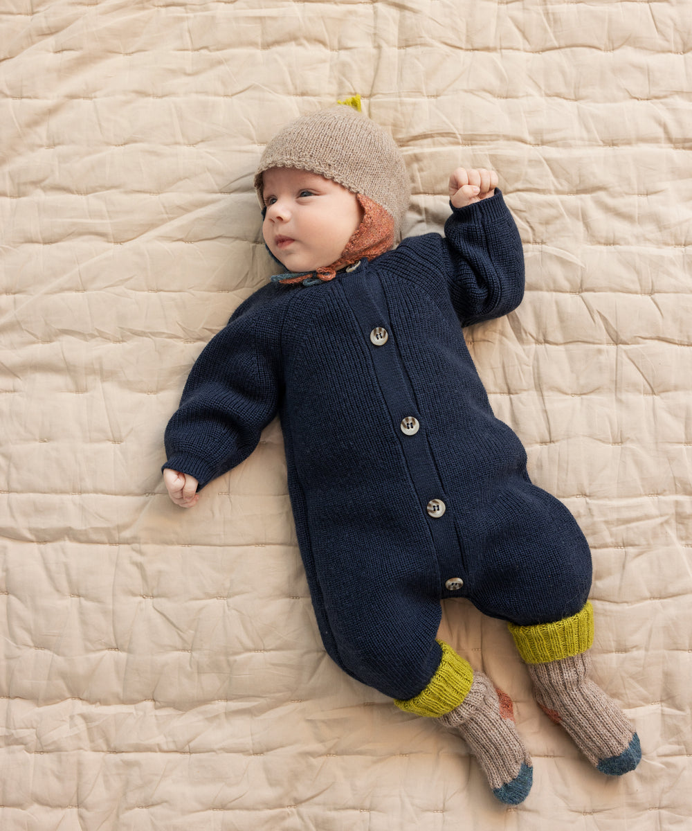 Baby ligger på tæppe iført beige uldhue og mørk heldragt