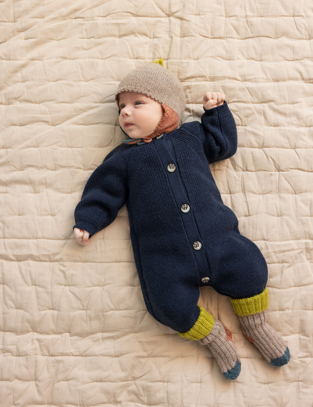 Baby ligger på tæppe iført beige uldhue og mørk holdragt.