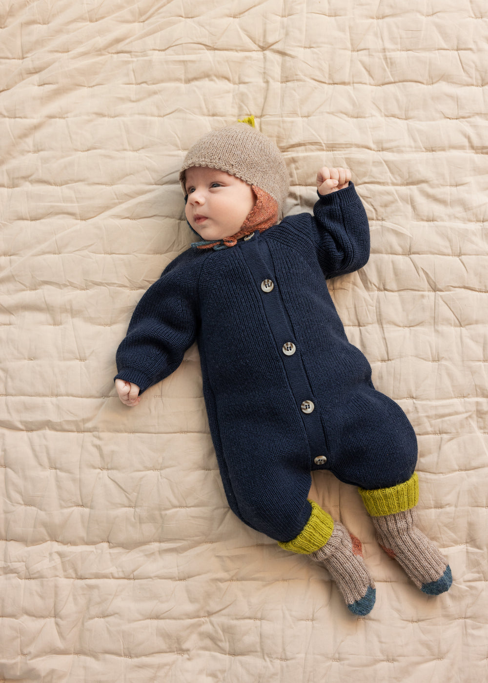 Baby ligger på tæppe iført beige uldhue og mørk heldragt