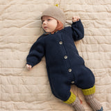 Baby ligger på tæppe iført beige uldhue og mørk heldragt