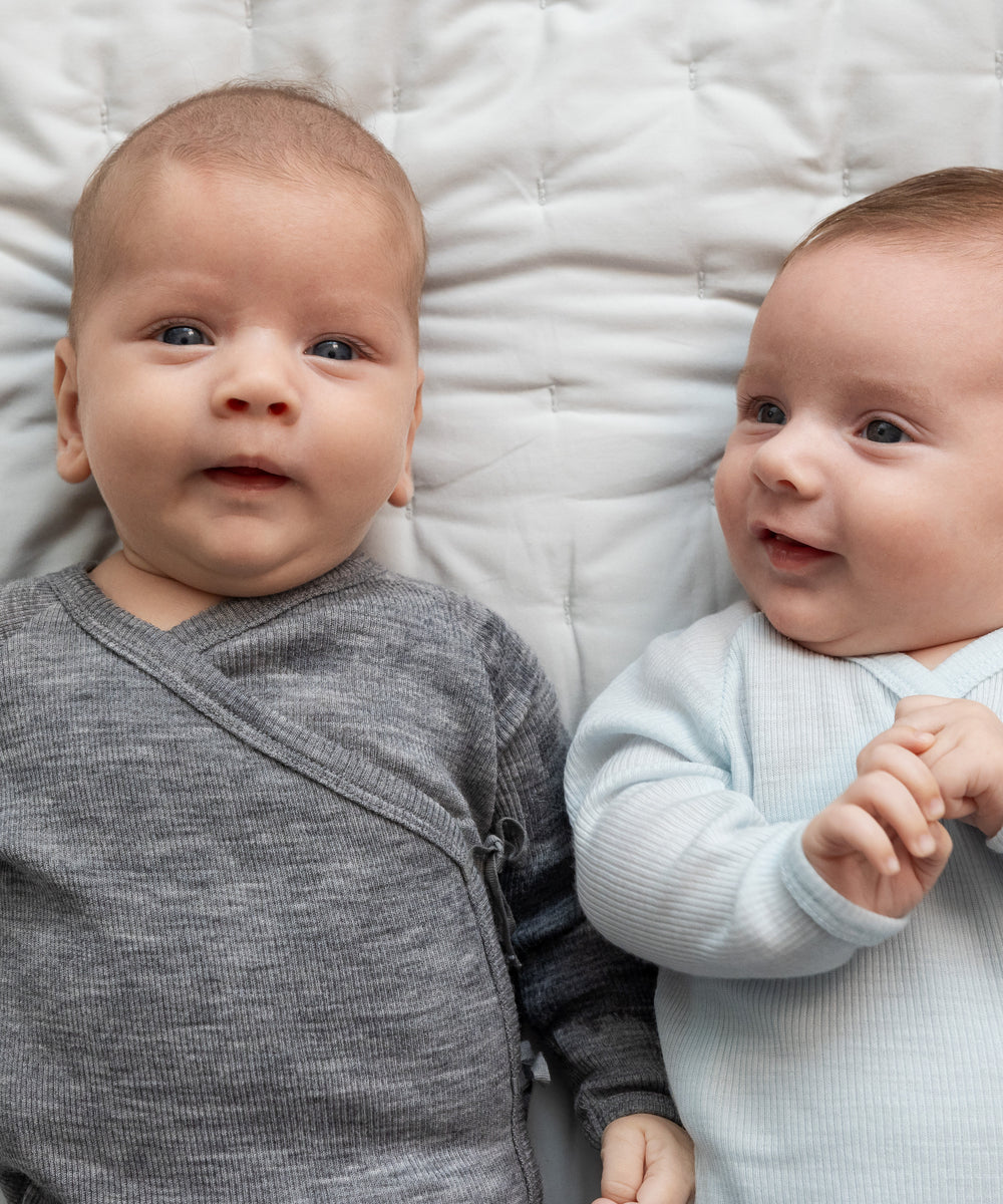 To babyer ligger på seng iført heldragter i merinould, en grå og en lyseblå