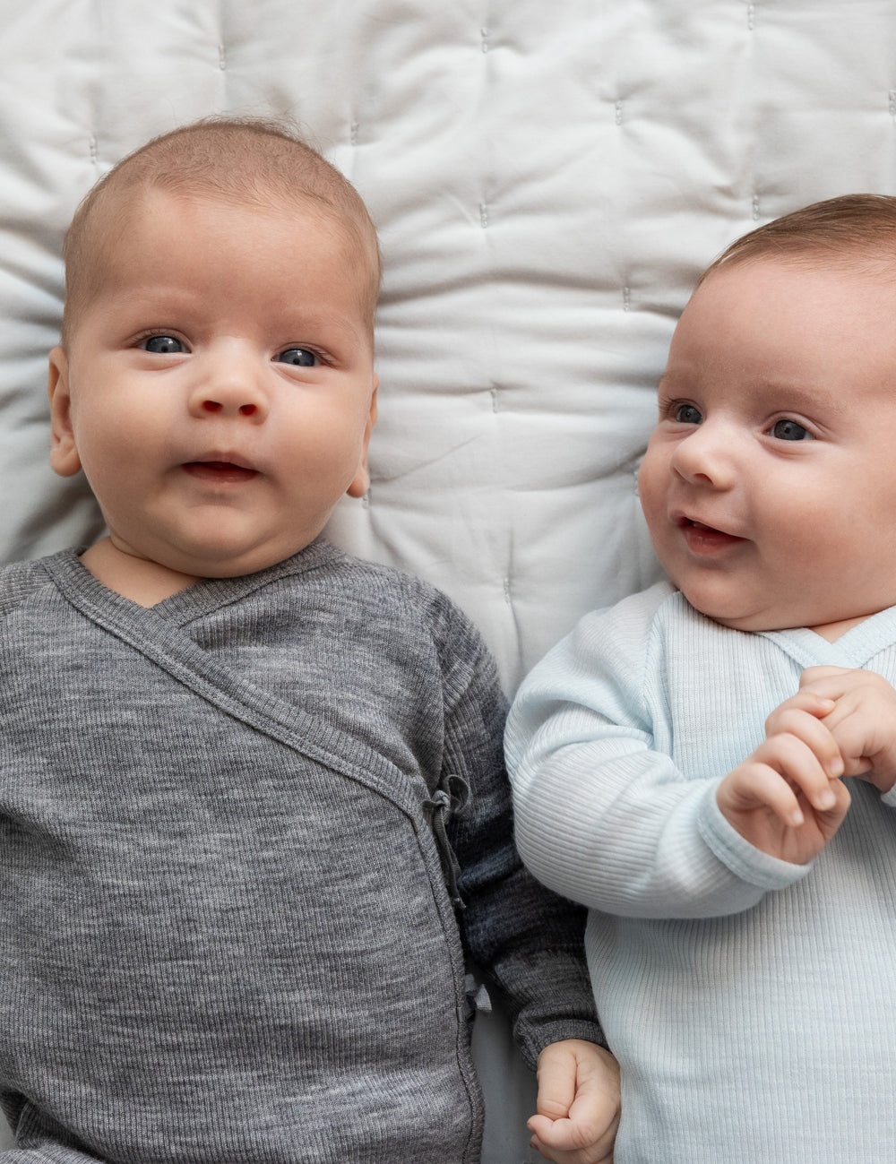 Bebåen ligger på seng iført holdragter i merino, en grå og en lyseblå.