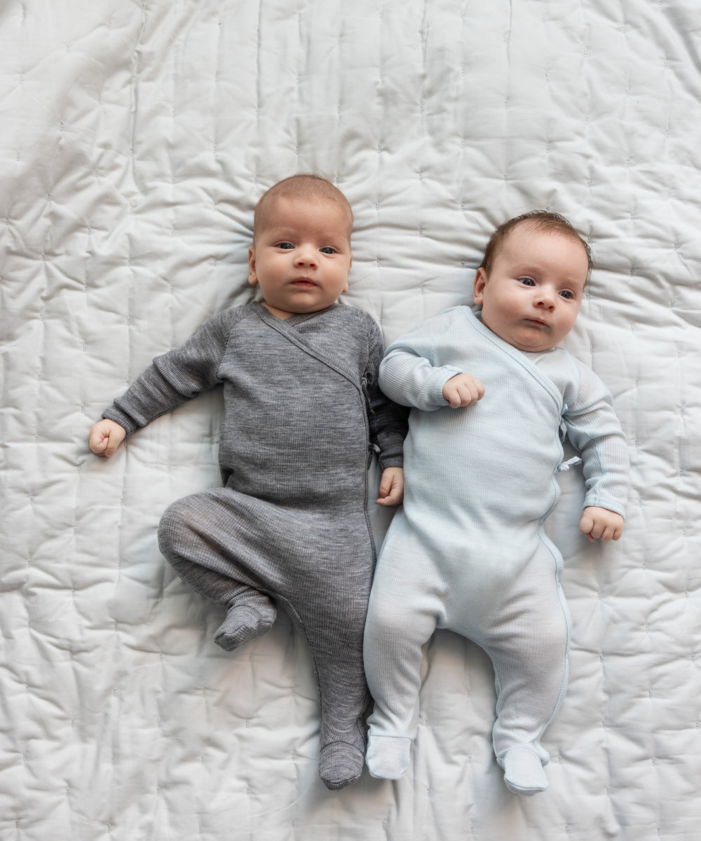 To babyer ligger på seng iført heldragter i merinould, en grå og en lyseblå