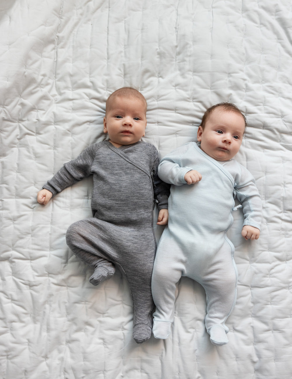 Bebåen ligger på seng iført holdragter i merino, en grå og en lyseblå.