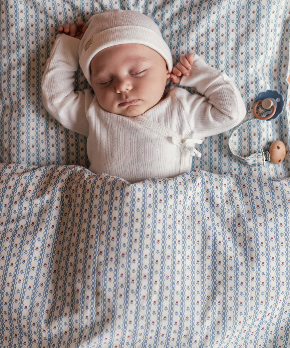 Baby iført hvid pointelle slå-om body og hue sovende under tæppe