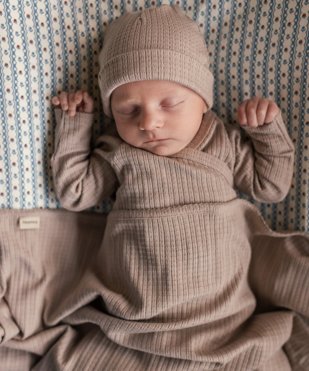 Baby iført beige pointelle sparkedragt og hue liggende med tæppe