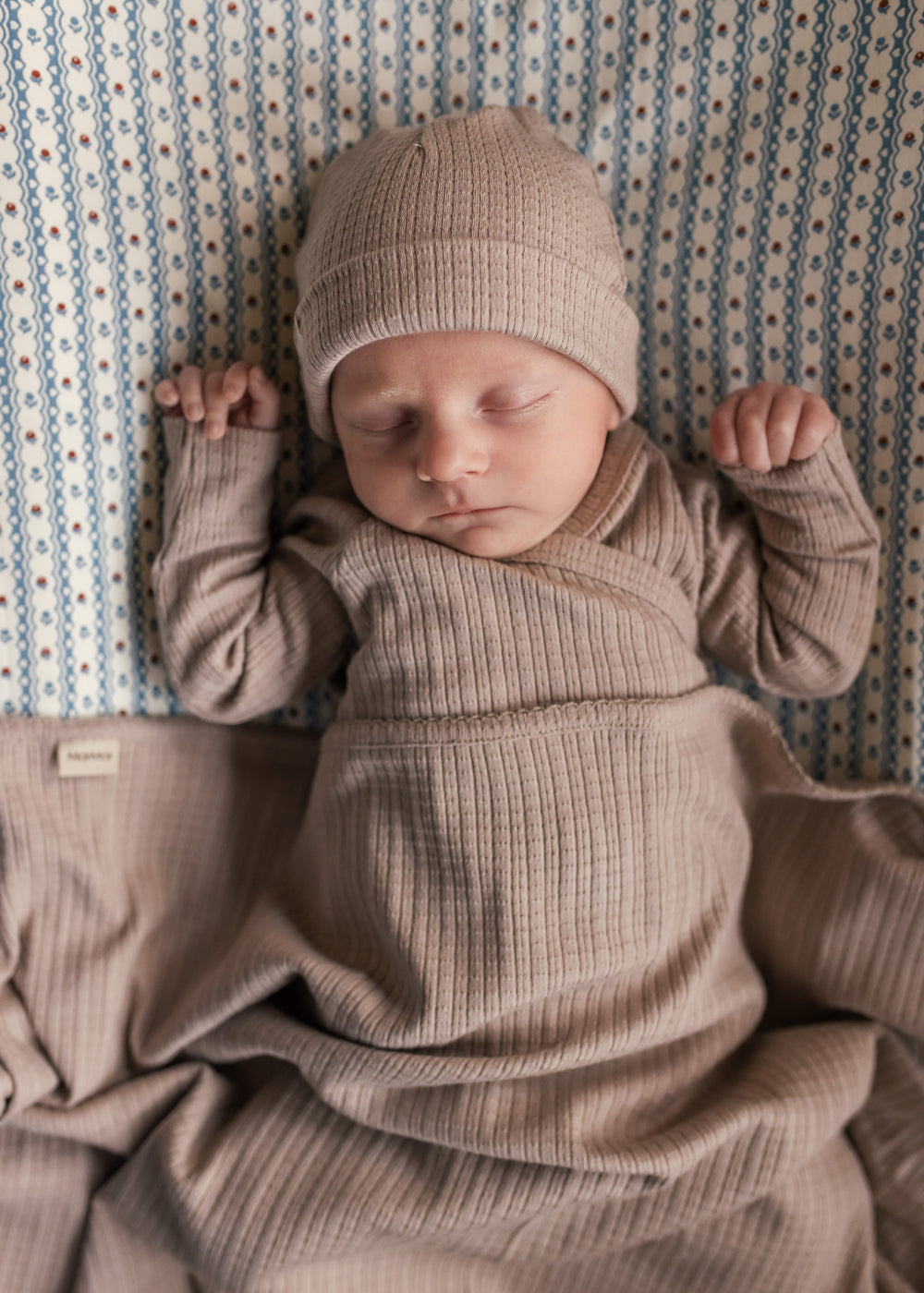 Baby iført beige pointelle sparkedragt og hue liggende med tæppe