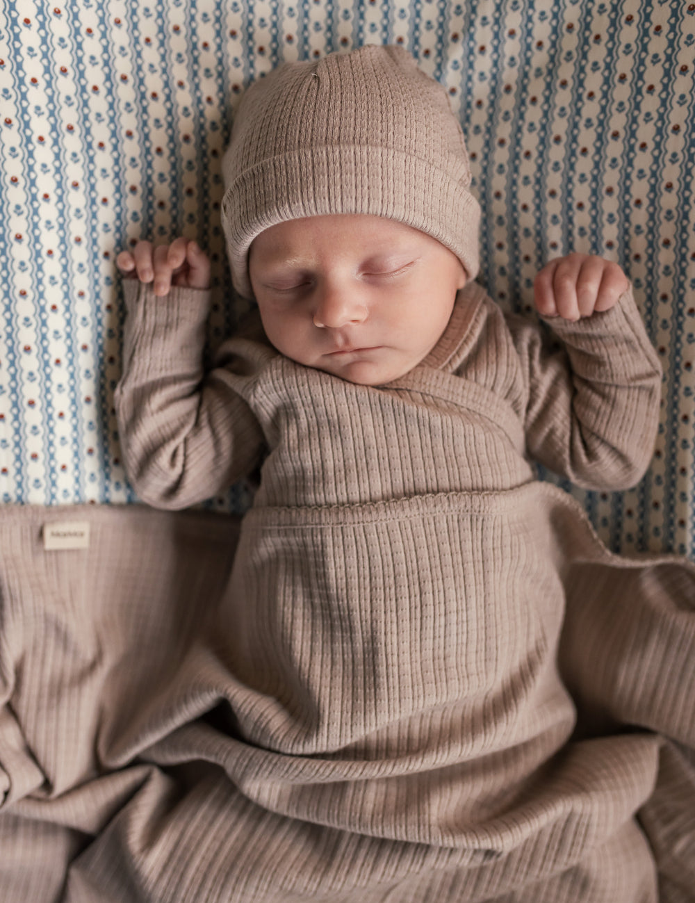 Baby iført beige pointelle sparkedragt og hue liggende på tæppe