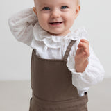 Lille børn iført hvid bluse med flæsekant og brune selebukser