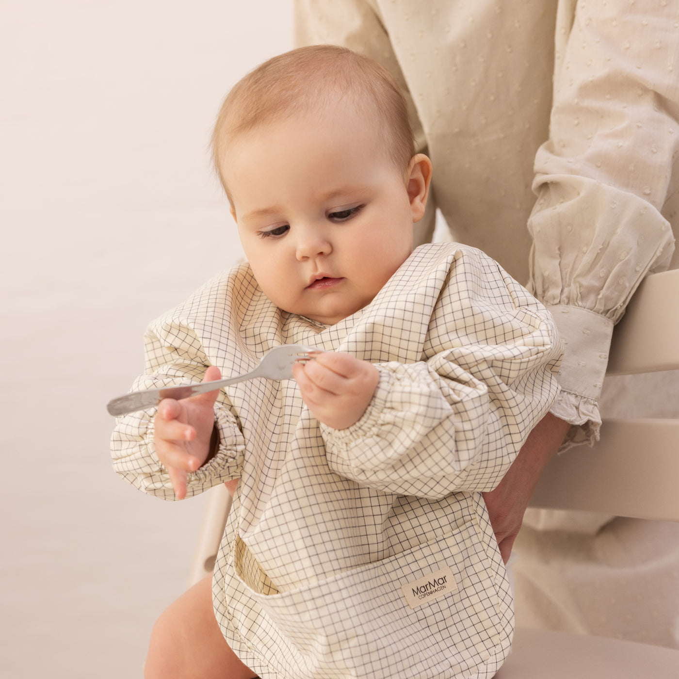 Baby iført ternet hagesmæk sidder og holder en gaffel