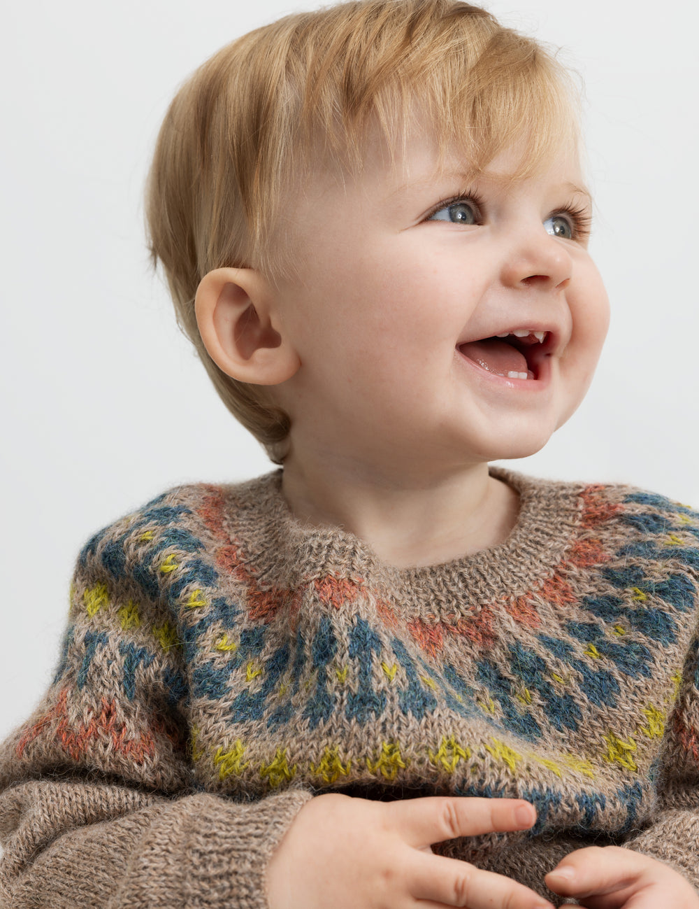 Baby smiler iført beige uldstrikbluse med farvet bærestykke