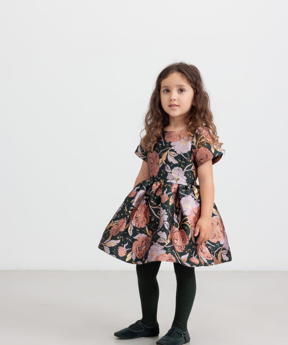 Pige iført mørkegrøn festkjole med blomsterprint og sorte strømpebukser til børn