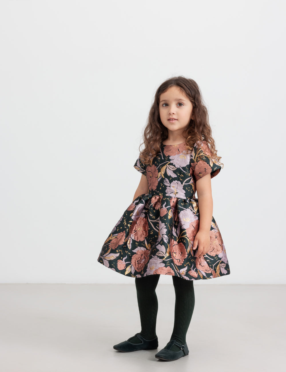 Pige iført mørkegrøn festkjole med blomsterprint og sorte strømpebukser til børn