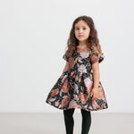 Pige iført mørkegrøn festkjole med blomsterprint og sorte strømpebukser til børn