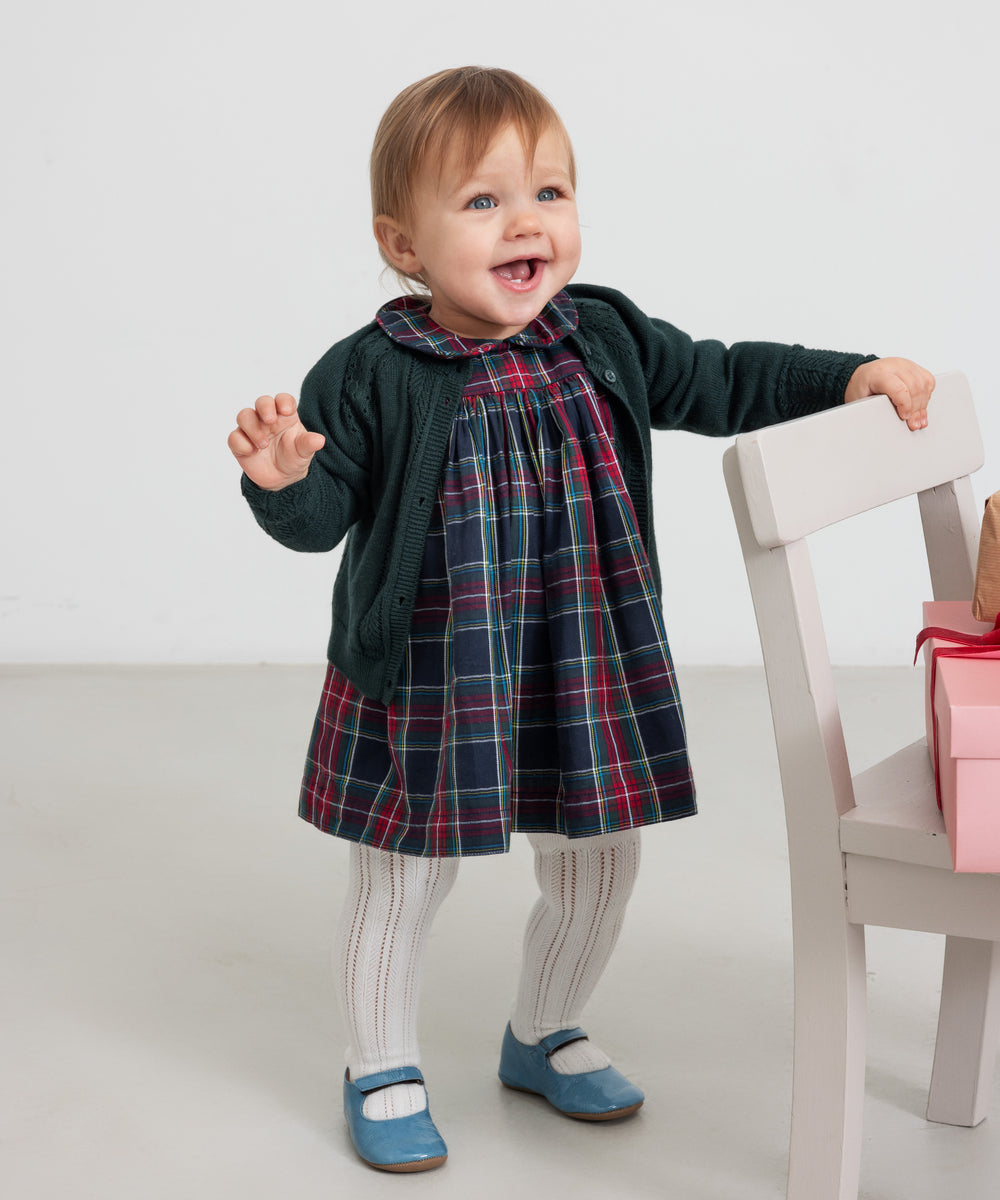 Baby iført mørkegrøn cardigan med hulmønster og ternet rød og grøn julekjole