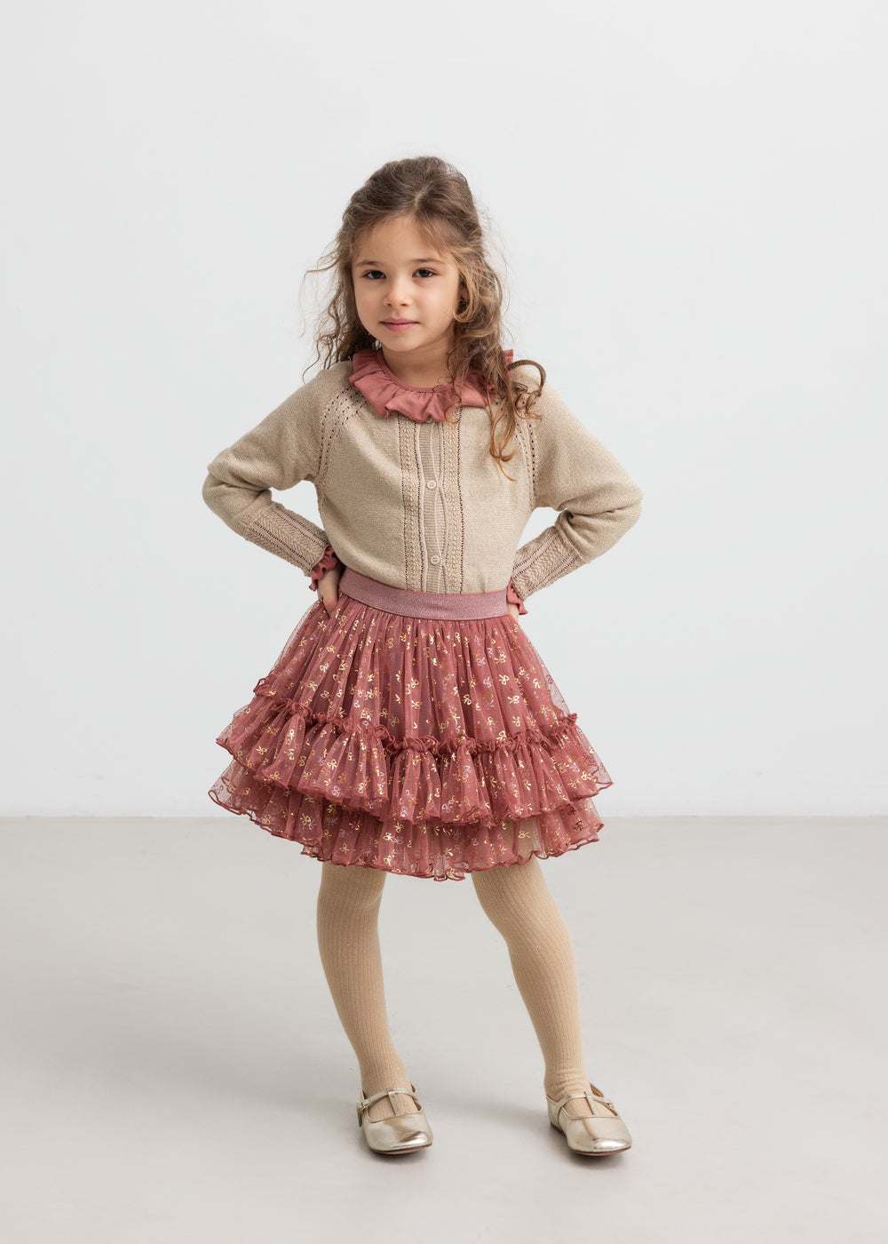 Pige iført beige cardigan med guldglimmer, lyserød festnederdel og strømpebukser til børn
