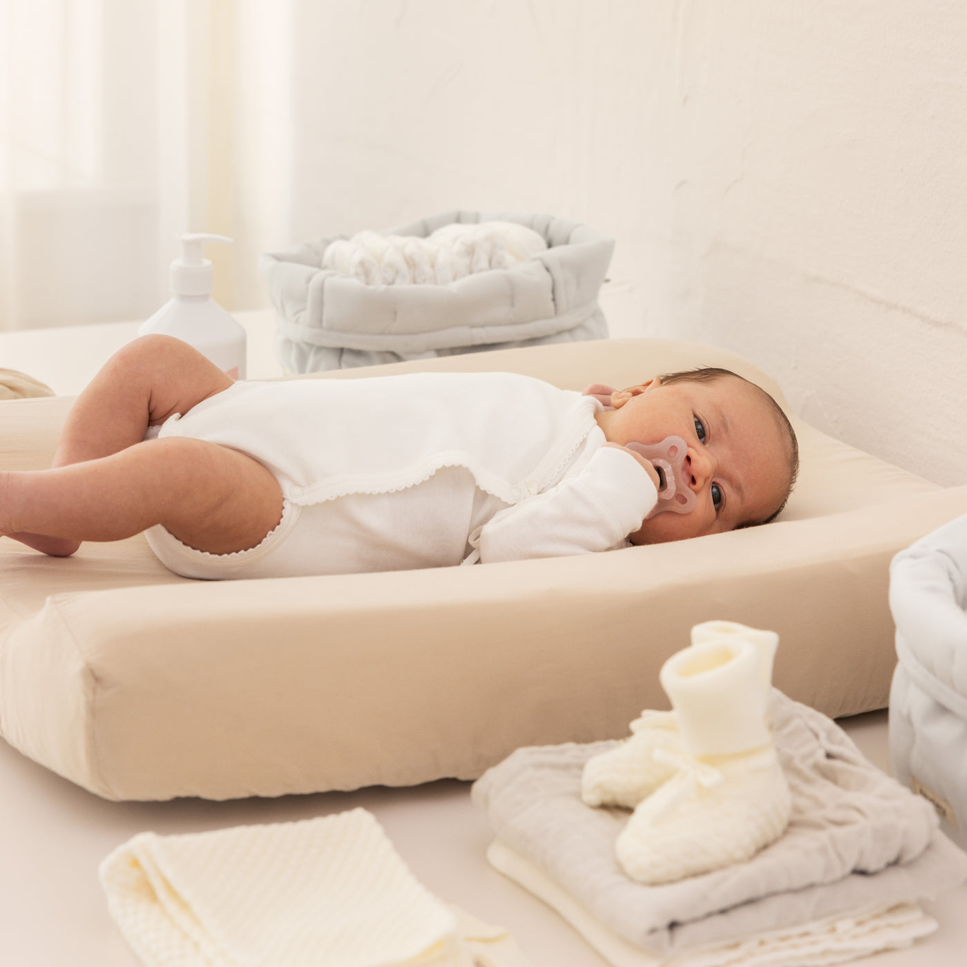 Baby i hvid body ligger på puslepude med sutter og små luffer ved siden af