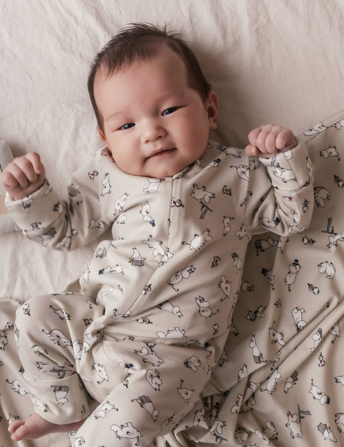 Nyfødt baby i beige heldragt med Moomin print liggende på seng med babytæppe