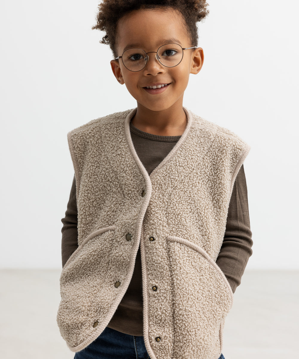 Barn iført lys teddyvest med hænderne i lommerne og brun t-shirt under