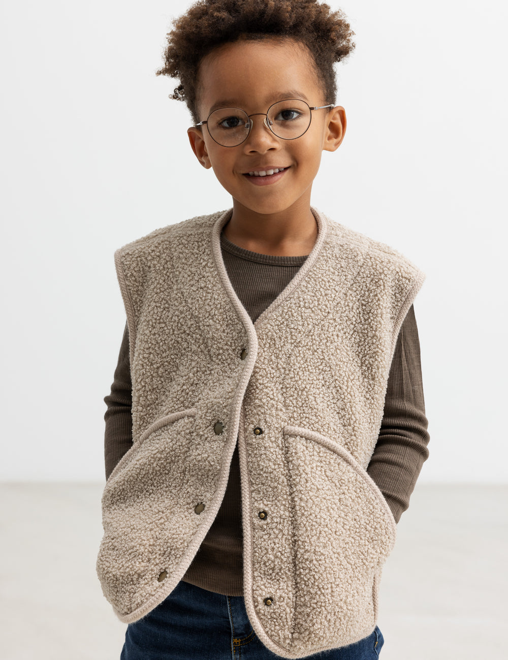 Barn iført lys teddyvest med hænderne i lommerne og brun t-shirt under.