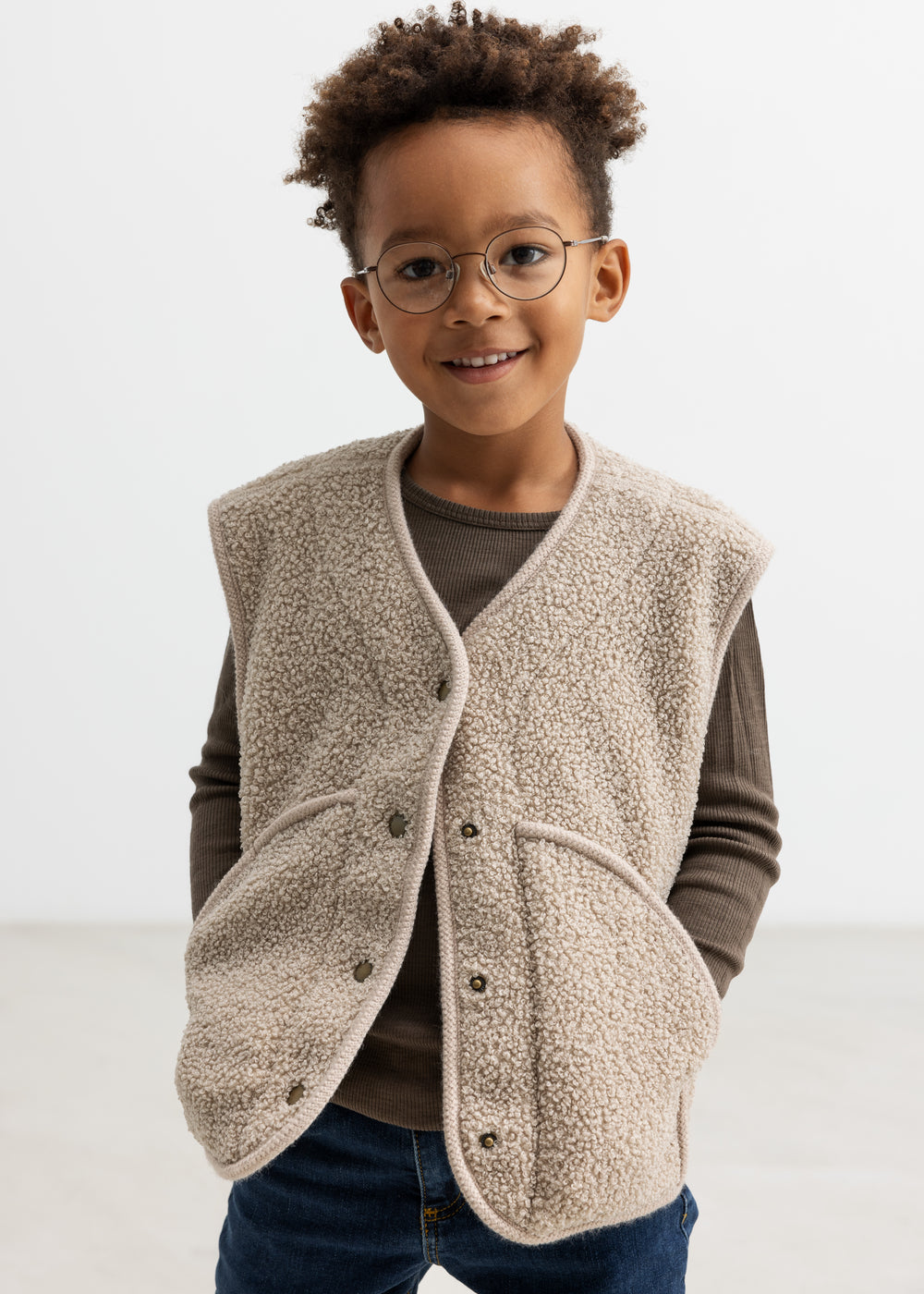 Barn iført lys teddyvest med hænderne i lommerne og brun t-shirt under