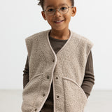 Barn iført lys teddyvest med hænderne i lommerne og brun t-shirt under