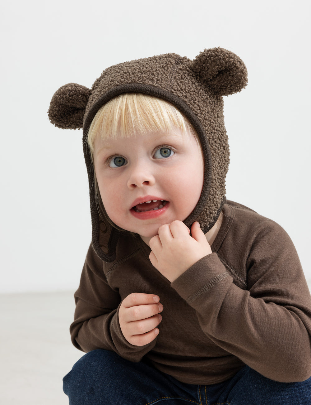 Barn iført brun elefanthue med ører i teddyfleece, sidder og ser op.