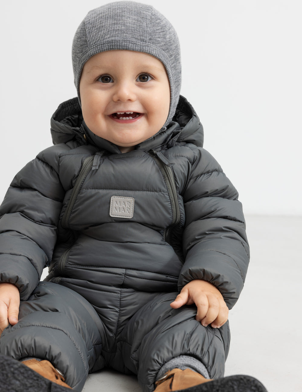 Smilende baby i mørkegrå dundragt sidder på gulv.