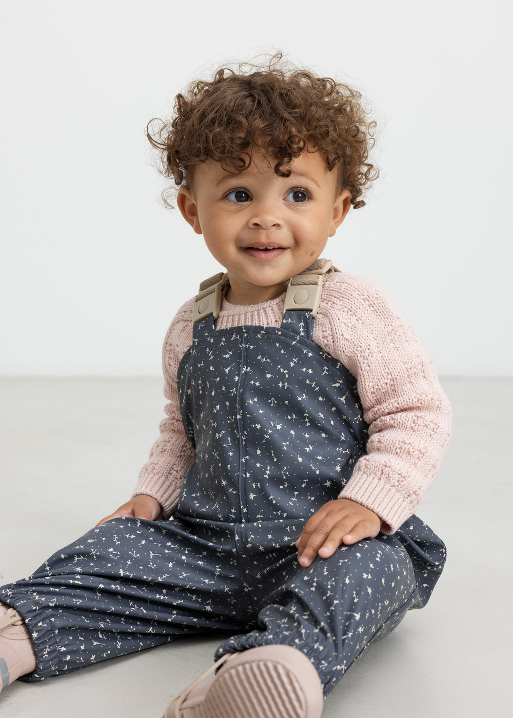 Barn iført mørkeblå regnbukser med print og lyserød trøje, sidder ned