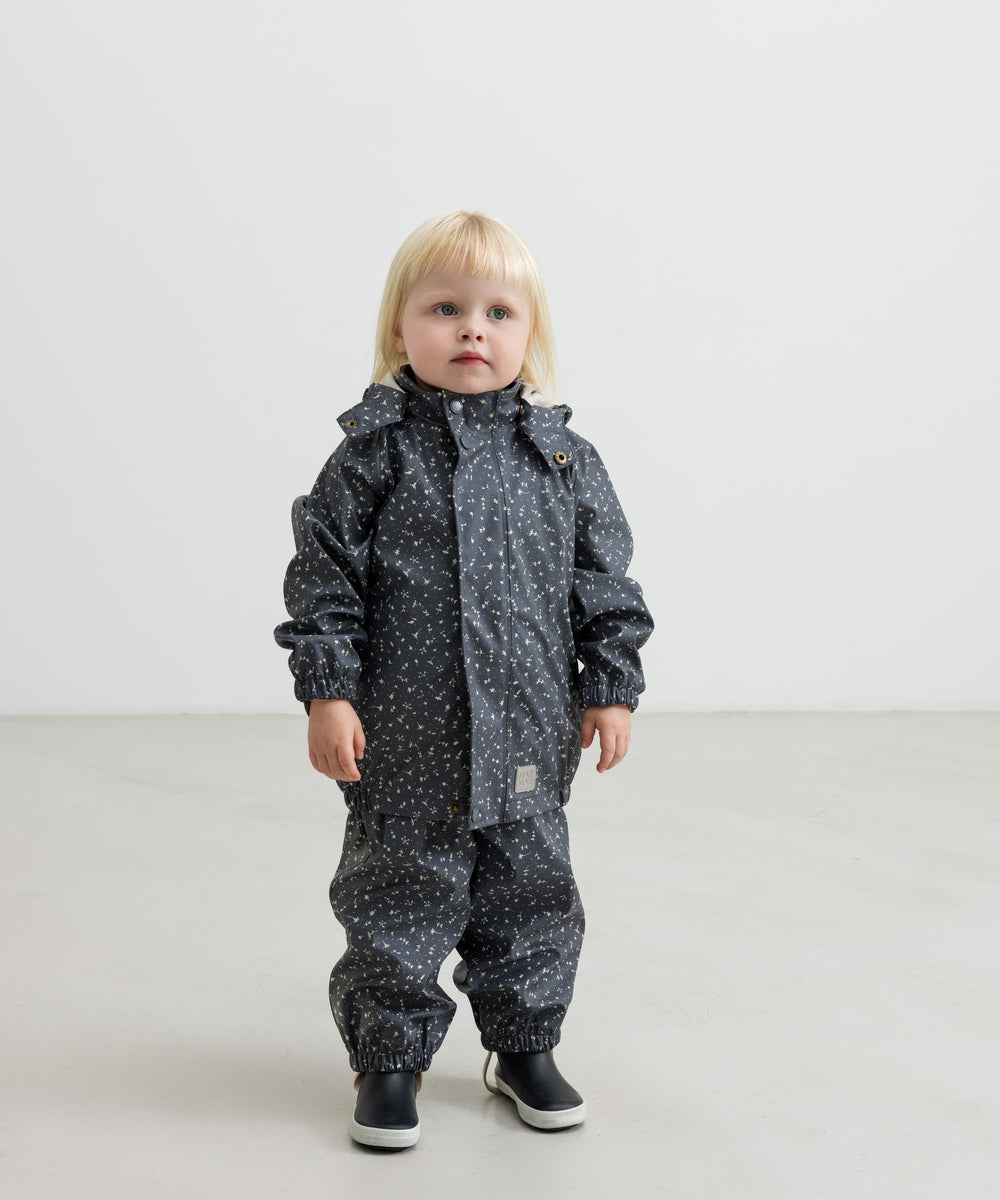 Barn i mørkeblå regnjakke med print og sorte støvler, står rakt op