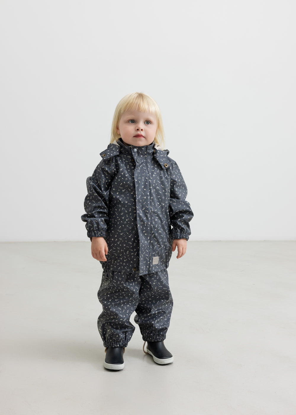 Barn i mørkeblå regnjakke med print og sorte støvler, står rakt op