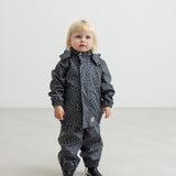 Barn i mørkeblå regnjakke med print og sorte støvler, står rakt op