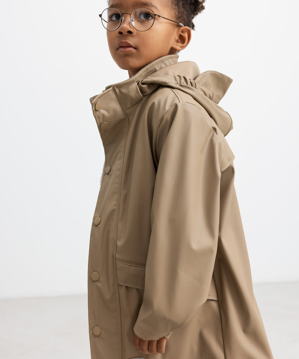 Barn iført beige regnjakke, set i profil med hætten oppe