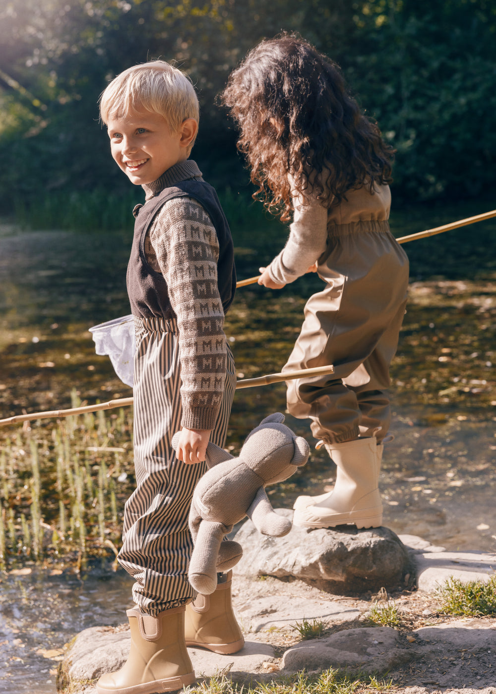 To børn leger ved vand i naturen iført regntøj, et med striber