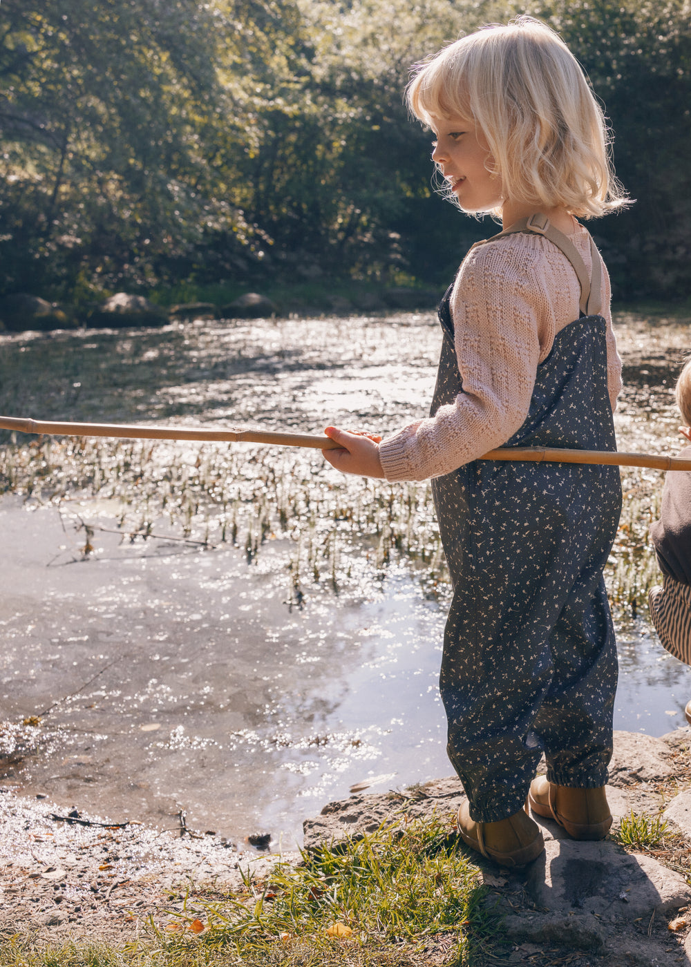 Børn iført mørkegrå regnbukser med prikker ved sø