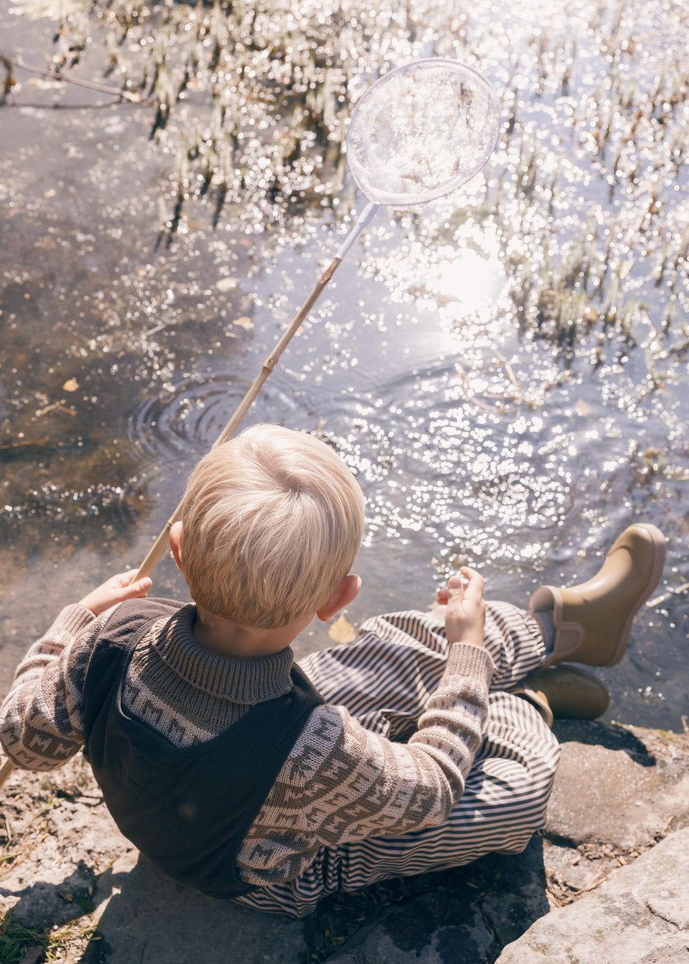 Dreng sidder ved sø iført brune stribede regnbukser
