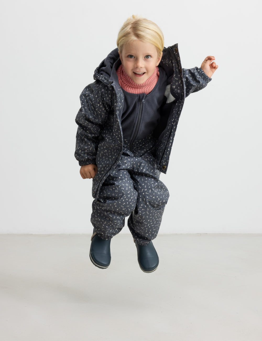 Børn hopper iført mørkegråt regnsæt med prikker og lyserød trøje.