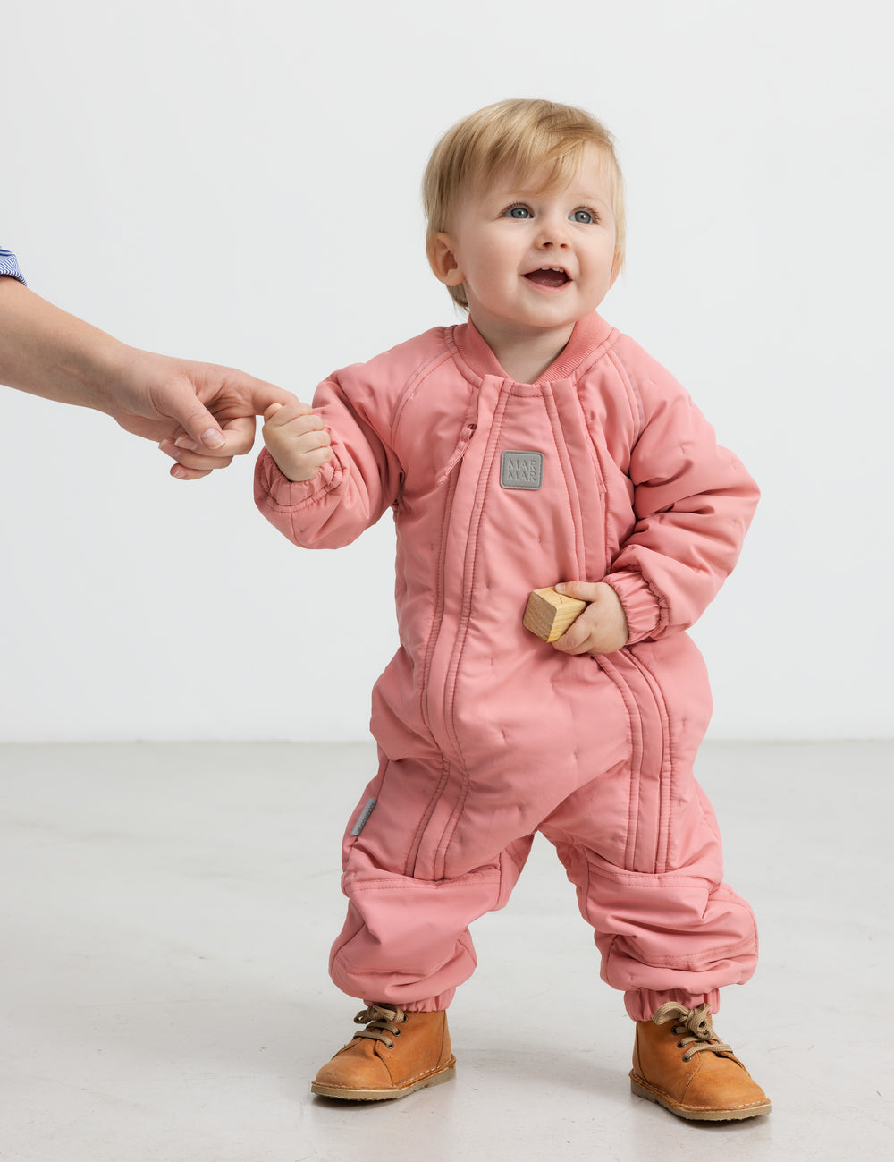 Baby iført lyserød termodragt, holder en voksen i hånden og smiler.
