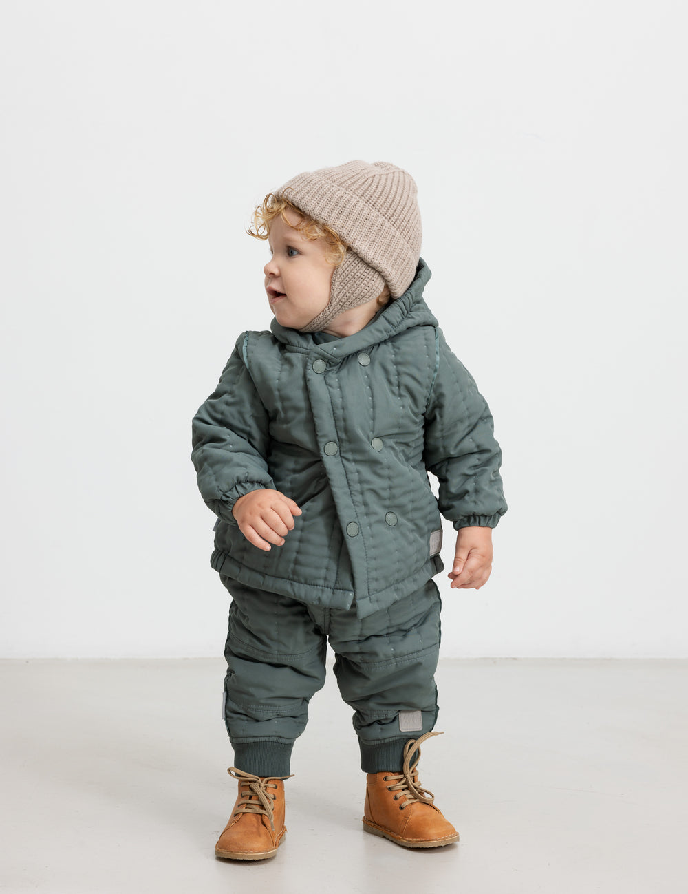 Baby iført støvet grøn termojakke og bukser, står og kigger til siden.