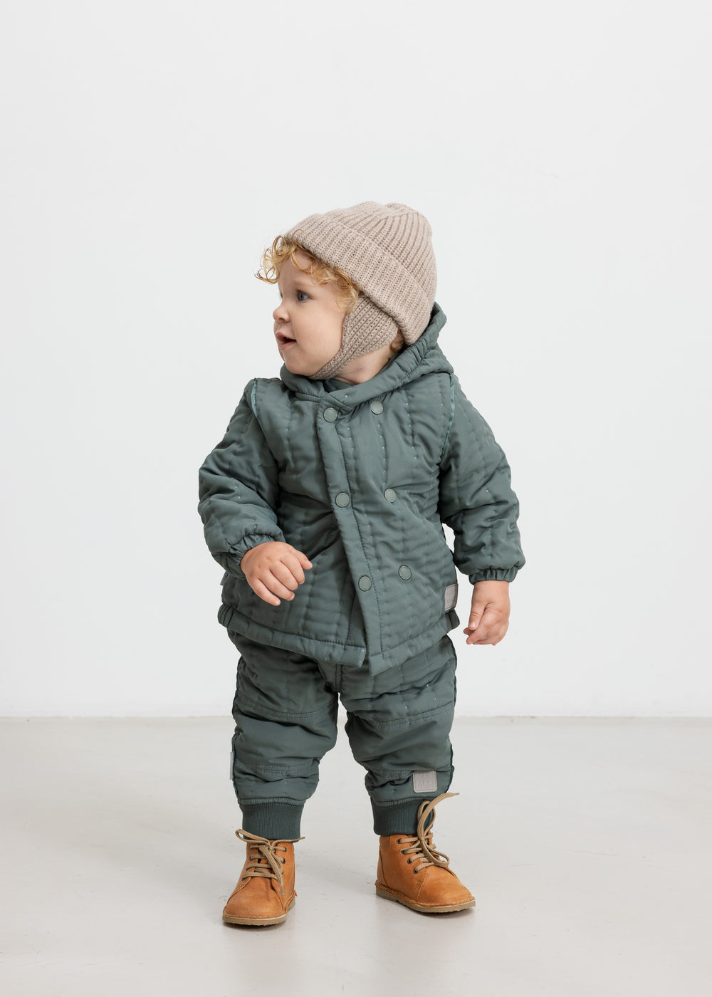 Baby iført støvet grøn termojakke og bukser, står og kigger til siden